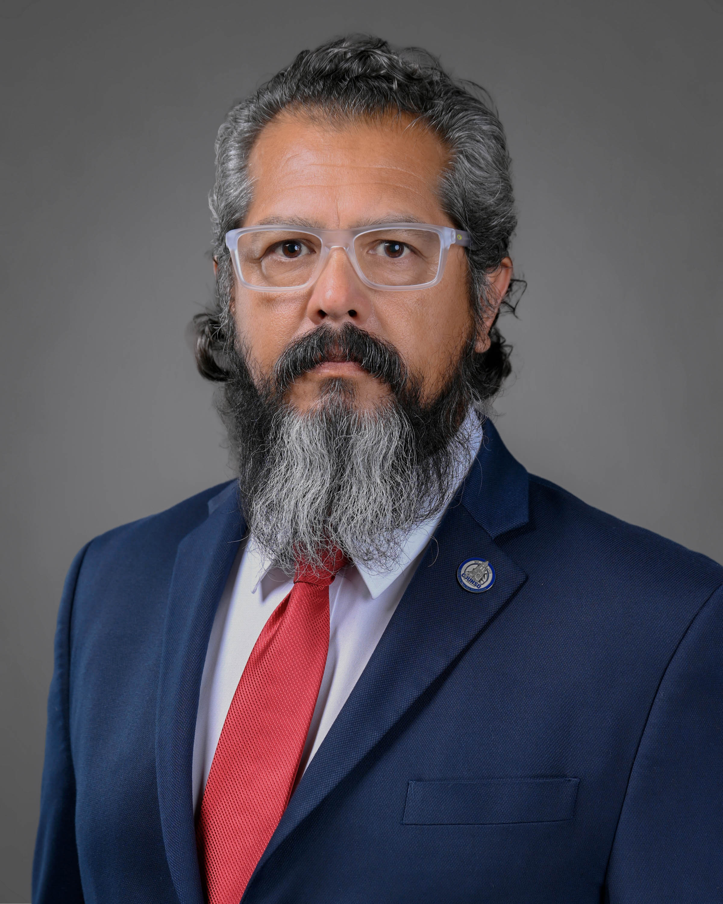 Professional headshot of a man with glasses and a long gray beard, wearing a navy suit, white dress shirt, and red tie against a gray studio background.